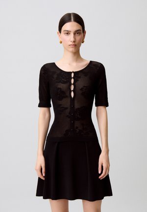 Mujer con el cabello peinado hacia atrás, lleva pendientes de aro dorados, una blusa negra ceñida con botones y estampado floral, y una falda negra de corte en línea A.