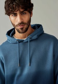 Donkerblauwe hoodie van zachte stof, met een buidelzak, trekkoordjes met metalen uiteinden en een gezellige capuchon. Eenvoudig ontwerp.