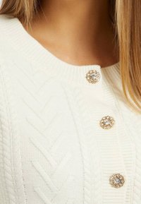 Biały sweter z dzianiny z warkoczowymi wzorami, okrągłymi kryształowymi guzikami z złotymi akcentami oraz ściągaczem przy dekolcie. Gładka tekstura.