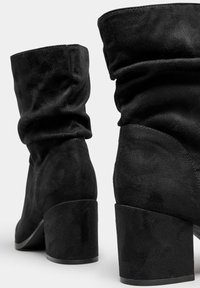 Botas de tobillo de ante negro con parte superior plisada, que cuentan con un tacón grueso y una textura suave y mate. Diseño sencillo sin adornos.