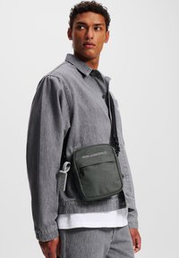 Zaino verde con tracolla regolabile, tasca a toppa e logo impresso. Indossato sopra una giacca in denim grigia e una camicia bianca. Design semplice e rettangolare.