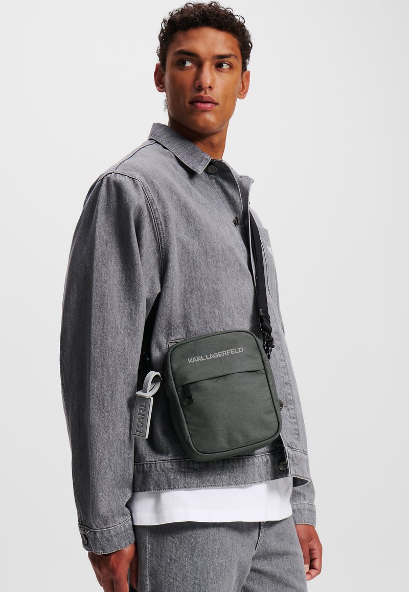 Zaino verde con tracolla regolabile, tasca a toppa e logo impresso. Indossato sopra una giacca in denim grigia e una camicia bianca. Design semplice e rettangolare.