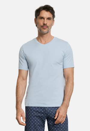 Mężczyzna ubrany w jasnoniebieską koszulkę z krótkim rękawem i dekoltem w serek oraz granatowe spodnie w wzory, stojący na jednolitym białym tle.