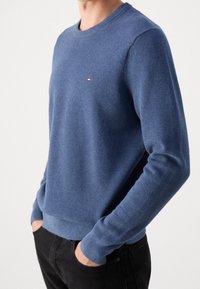 Maglione blu a collo rotondo con una trama testurizzata, polsini e orlo a costine, caratterizzato da un piccolo logo sul petto. Abbinato a pantaloni neri.