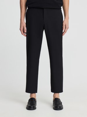 Osoba ubrana w czarne, krótkie spodnie o prostych nogawkach i czarne loafersy, stojąca na tle jednolitego, jasnego tła.