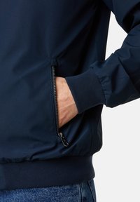Mano inserita nella tasca frontale con zip di una giacca blu navy con polsini e orlo a costine, abbinata a jeans blu.