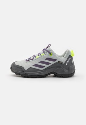 Chaussure de randonnée gris clair et gris foncé avec des rayures violettes, des lacets violets, des œillets jaune néon et une semelle noire texturée sur un fond neutre.
