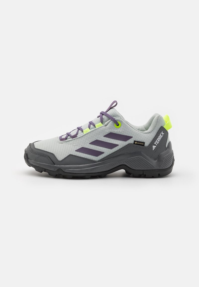 Chaussure de randonnée gris clair et gris foncé avec des rayures violettes, des lacets violets, des œillets jaune néon et une semelle noire texturée sur un fond neutre.