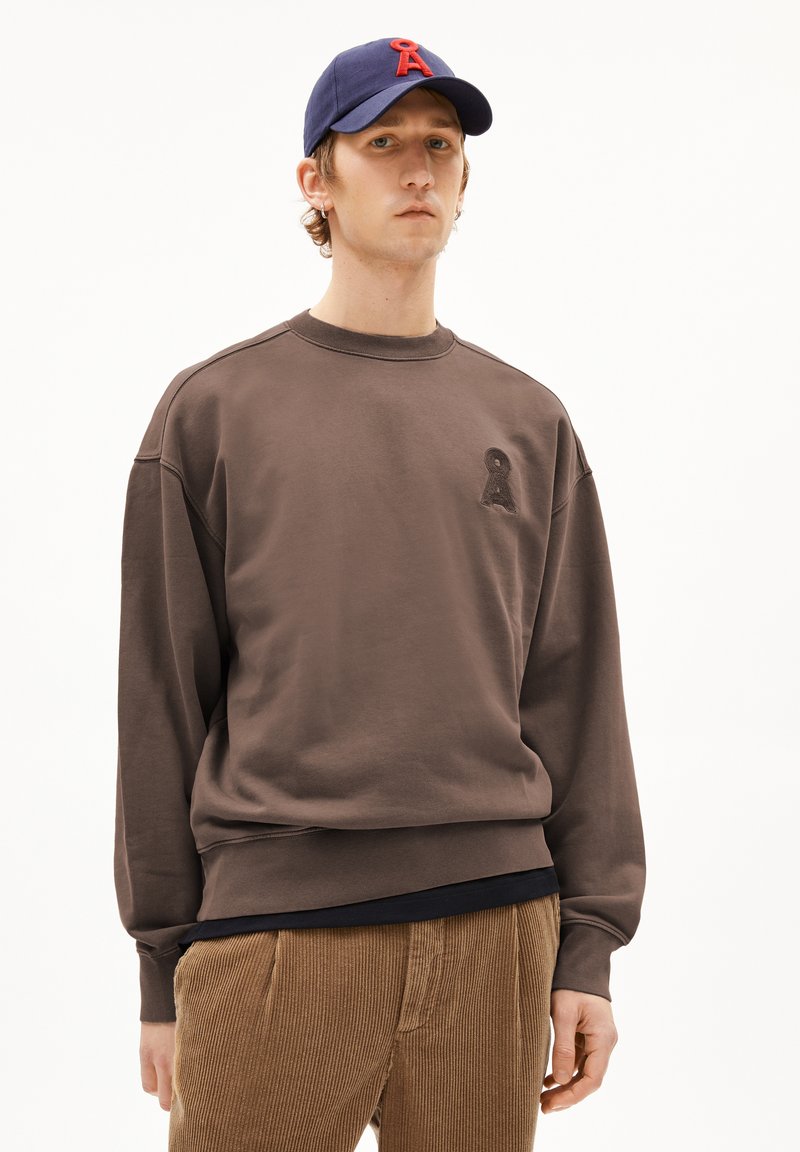 Bruine sweatshirt met drop shoulders en geribbelde mouwen, met een klein zwart logo op de borst. Gecombineerd met tan corduroy broek.