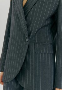 Mørkegrå blazer med striber, med klapkrave, enkelt knaplukning og en brystlomme; stoffet har en glat tekstur.