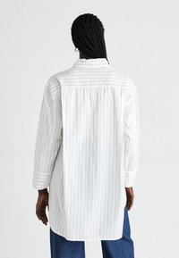 Persona con capelli intrecciati, che indossa una lunga camicia bianca a righe sottili e jeans blu, in piedi di spalle contro uno sfondo semplice.