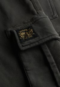 Tessuto grigio scuro, con una superficie strutturata. È visibile un'etichetta nera con testo dorato che indica il marchio "Superdry" e i dettagli.