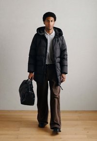 Casaco acolchoado preto com capuz, camisa polo cinza, calças largas castanhas e mala preta texturizada. Design simples sobre um piso de madeira clara.