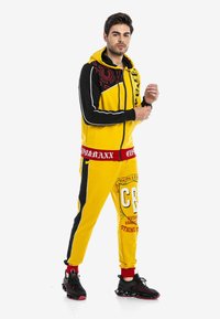 Helle gelbe Trainingsanzug mit schwarzen Akzenten, rotem Bund mit Logo und Grafikdesign auf den Hosen. Trägt schwarze Sneakers.
