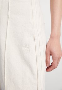 Närbild av beige Adidas-byxor med tre vertikala ränder och en broderad logo nära höften, tillsammans med en avslappnad hand vid sidan.