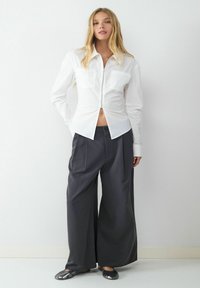 Weiße Reißverschlussbluse mit zwei Brusttaschen, kombiniert mit weit geschnittenen, dunkelgrauen Hosen. Weicher Stoff, minimalistisches Design und glatte Textur.