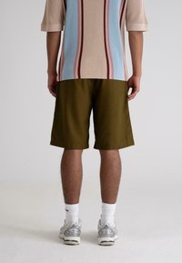 Oliven grønne shorts i bomull med avslappet passform, kombinert med en stripete teksturert skjorte i pastellfarger. Modellens bakside, stående mot en lys bakgrunn.