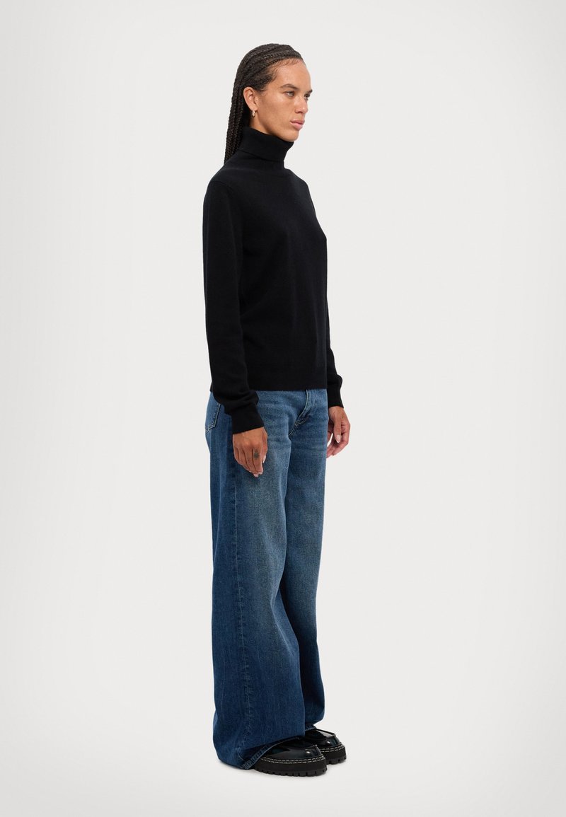 Black turtleneck sweater and wide-leg blue jeans, with a smooth texture. Model stands sideways, wearing black shoes with a chunky sole.