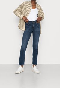 Camisa de cuadros marrón claro sobre una camiseta blanca de cuello en V, combinada con jeans ajustados azul oscuro y zapatillas blancas. Diseño simple con líneas limpias.