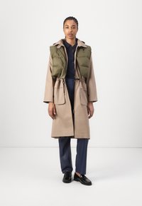 Manteau long beige avec taille à cordon de serrage, surmonté d'un gilet rembourré vert. Chemise et pantalon bleu marine, chaussures noires. Design minimaliste.