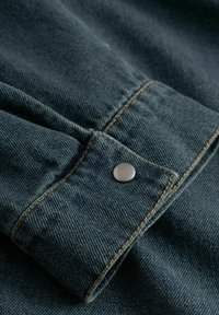 Țesătură din denim albastru închis cu cusături portocalii și nasture metalic argintiu pe un manșon pliat.