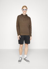 Felpa marrone con cappuccio e tasca frontale, abbinata a pantalonicini cargo neri. Sneakers grigie e occhiali da sole blu completano l'outfit.