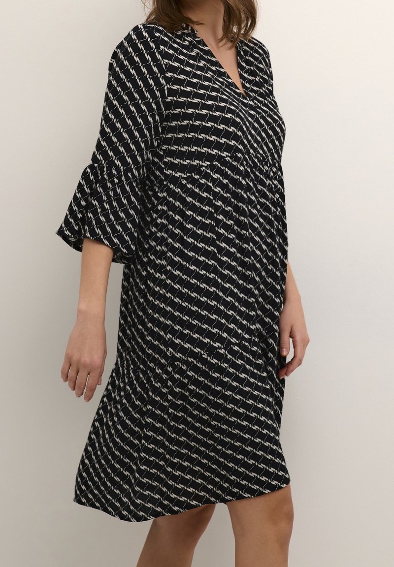 Robe noire à motif géométrique blanc, col en V et manches à volants. Tissu fluide, longueur genou, coupe décontractée.