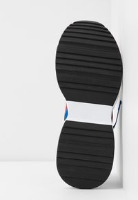 Suela de zapato de goma negra con ranuras horizontales, entresuela blanca con una sección de acento azul y rojo, colocada sobre una superficie blanca.