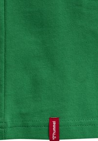 Grüner Stoff mit sichtbaren Nähten und einem kleinen roten Stoffetikett mit der Aufschrift "hummel" nahe dem unteren Saum.