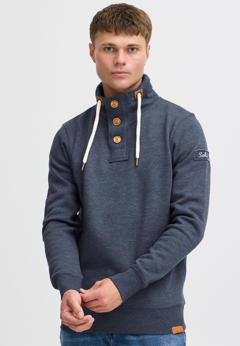 Donkerblauwe sweatshirt met een hoge kraag, houten knoopsluiting en crèmekleurige trekkoorden. Heeft ribstof manchetten en een klein logopatch op de mouw.