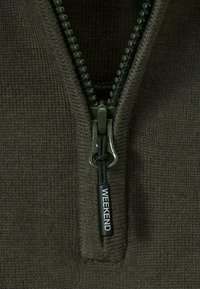 Lähikuva vihreästä vetoketjusta neulottu kankaalla, jossa on musta vetotappi, jossa lukee "WEEKEND" valkoisella tekstillä.