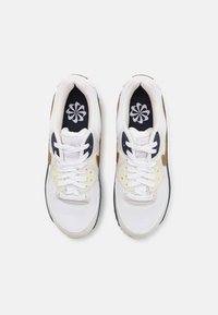 Par bijelih atletskih cipela s zlatnim i mornarsko plavim akcentima, bijelim vezicama i cvjetnim uzorkom na tamnim ulošcima, pregledano odgore.