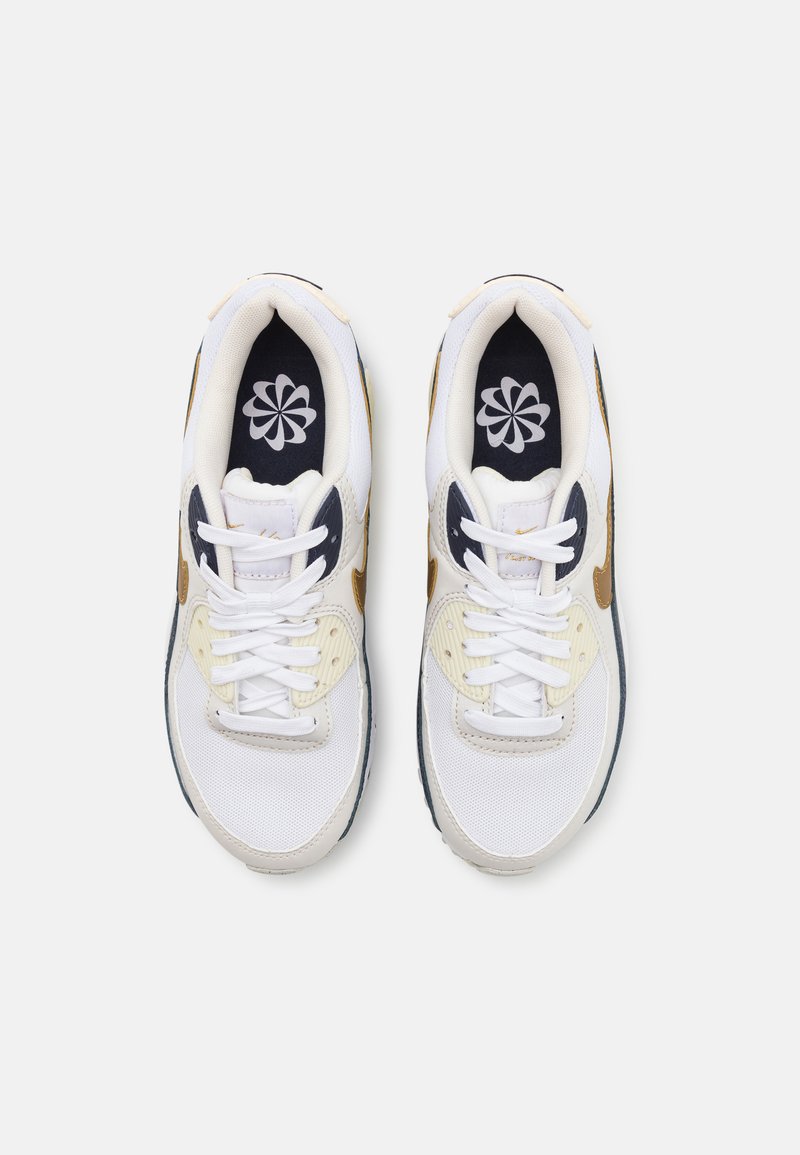 Un par de zapatillas deportivas blancas con acentos dorados y navy, cordones blancos y un patrón floral en las plantillas oscuras, vistas desde arriba.