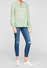 Blusa verde chiaro a maniche lunghe con scollatura rotonda e dettaglio con fiocco, abbinata a jeans skinny blu strappati e sneakers bianche.