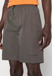 Shorts gris avec une taille élastique et un cordon de serrage, dotés de poches latérales et d'un logo imprimé près de l'ourlet. Texture lisse, design décontracté.