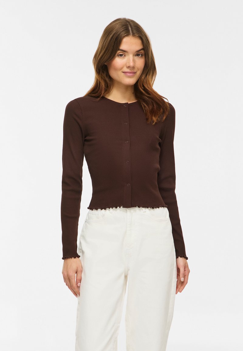 Jeune femme portant un haut marron ajusté à manches longues boutonné et un pantalon blanc taille haute, debout devant un fond uni.