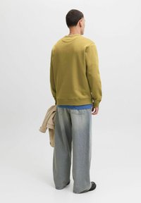 Felpa verde oliva con polsini e orlo a coste, abbinata a jeans a gamba larga in denim chiaro. Una giacca esterna beige è tenuta nella mano sinistra.