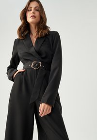 Zwarte jumpsuit met een wikkelontwerp, lange mouwen en een enkele borstzak. Gladde textuur en een metalen gespaccent.