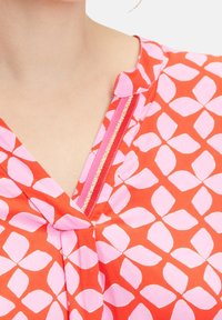 Gros plan sur une personne portant un haut à motifs rose et orange avec un décolleté en V et une finition à rayures dorées sur le col.