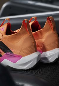 Paire de chaussures de sport orange avec semelles blanches, accents noirs et roses, et languettes au talon, placées sur une surface noire texturée.