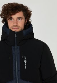 Svart puffjacka med hög krage, kontrasterande marinblå detaljer, framdragkedja och logotypdetalj. Tyget verkar slätt med en vadderad struktur.
