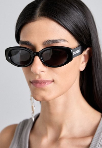 Lunettes de soleil rectangulaires noires avec verres foncés et branches épaisses ornées de texte blanc. Le modèle a les cheveux noirs et raides et porte un haut gris.
