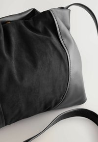 Geantă neagră din suede și piele, cu texturi netede, forme delicate, detalii subtile ale cusăturilor și o curea lungă și subțire pentru umăr.