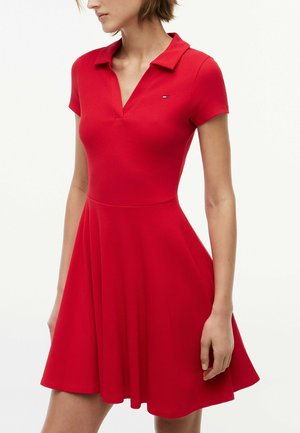 Frau trägt ein rot kurzärmeliges Polo-Kleid mit ausgestelltem Rock und kleinem Logo auf der Brust vor weißem Hintergrund.