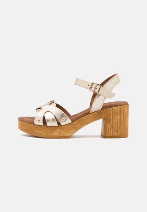 Sandalias metálicas doradas con tacón bloque de madera, con tiras cruzadas, un lazo decorativo y detalles de metal redondos en la parte superior.
