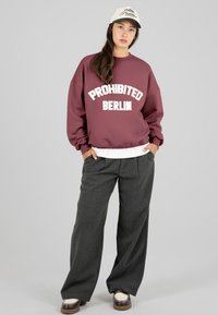 Žena stojící s rukama v kapsách, oblečená v vínovém svetru "PROHIBITED BERLIN", šedých volných kalhotách, bílé kšiltovce a mokasínech.