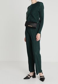 Haut plissé vert à manches longues associé à un pantalon assorti, sac bandoulière noir avec des accents dorés et chaussures noires pointues avec des brides.