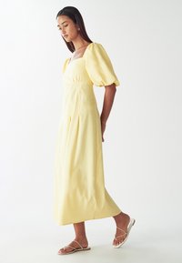 Žlté ľanové šaty s hranatým výstrihom a balónovými rukávmi, so vypasovaným pásom a splývavou sukňou. Nosené s nahými plochými sandálmi.