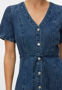 Denimist lühikeste varrukatega kleit, V-kaelusega, ees nööbiga kinnitatav ja ise siduvat vööd kandval inimesel paljaste kätega.