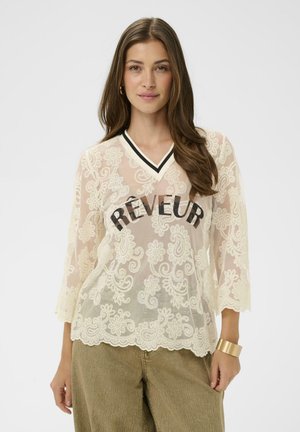 Jeune femme aux longs cheveux bruns portant un haut en dentelle blanche transparente avec le texte « RÊVEUR » et un pantalon vert olive, debout devant un fond uni.